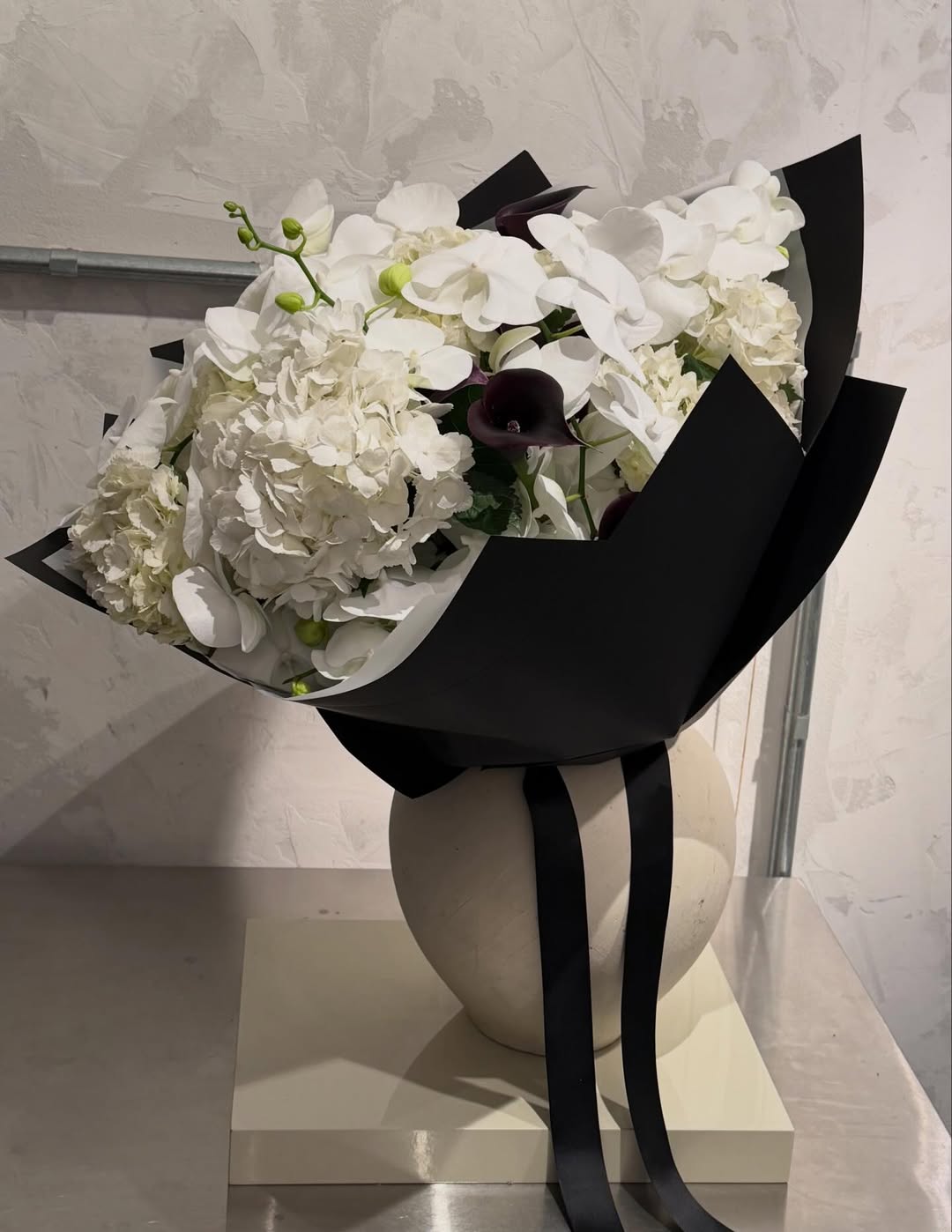 Buquê de hortênsias e orquídeas brancas com flores escuras, envolto em papel preto e fita preta.